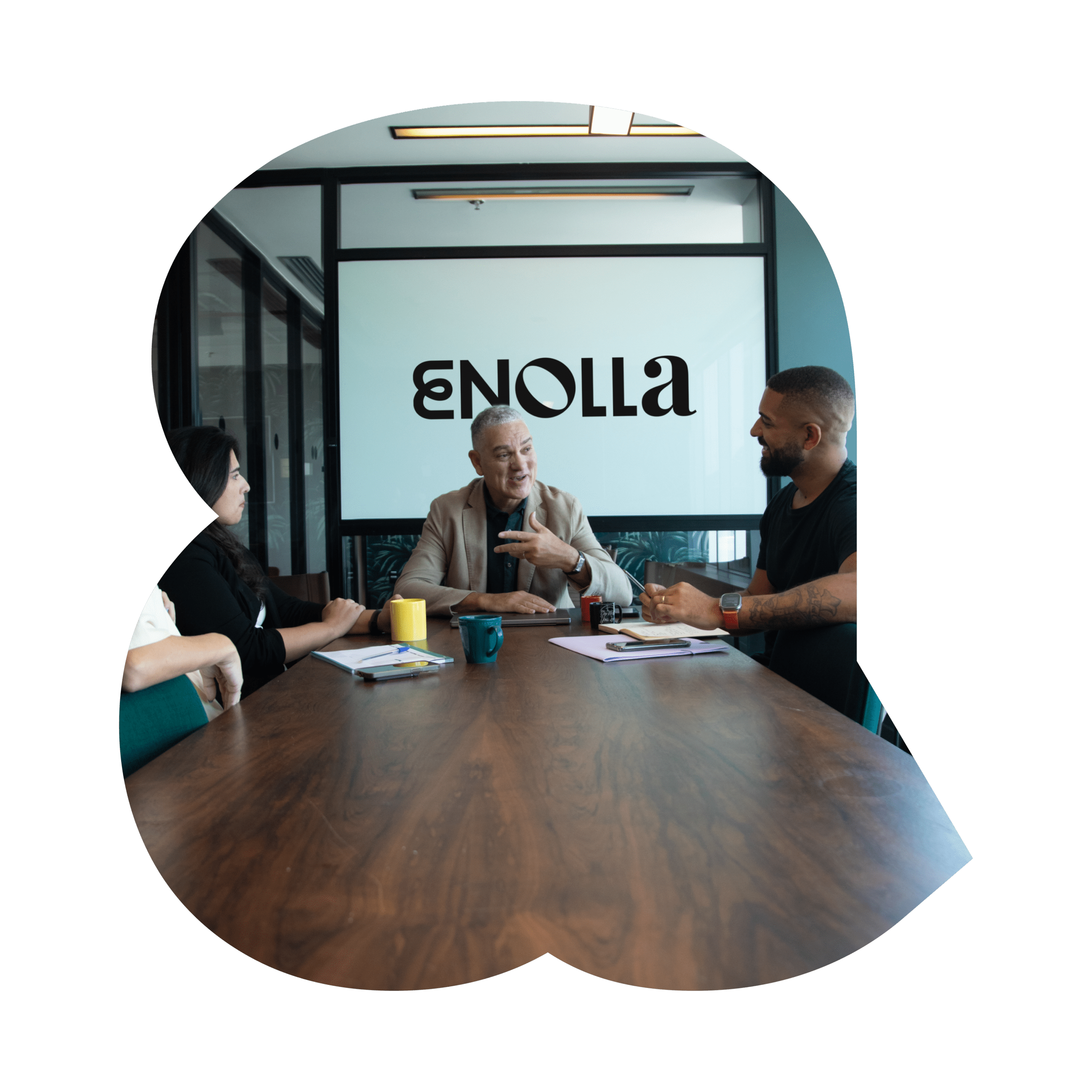 A group of four coworkers sit around a wooden conference table in a modern office meeting room, engaged in conversation. A man in the center gestures while speaking, behind them displays the word ‘ENOLLA.